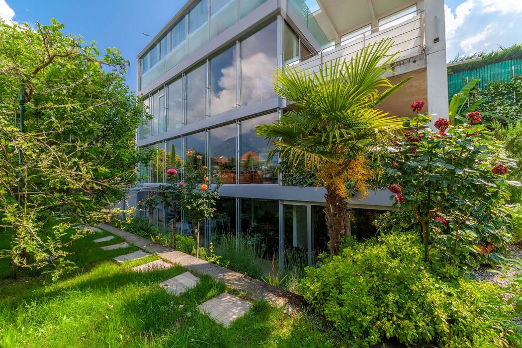 Modernes Glashaus mit üppigem Garten, Palmen und blühenden Rosen.