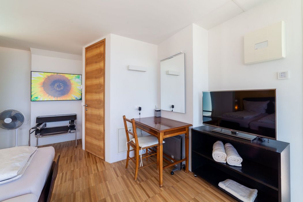 Helles Hotelzimmer mit Holzboden, Schreibtisch und Stuhl, TV und großem Sonnenblumenbild an der Wand.