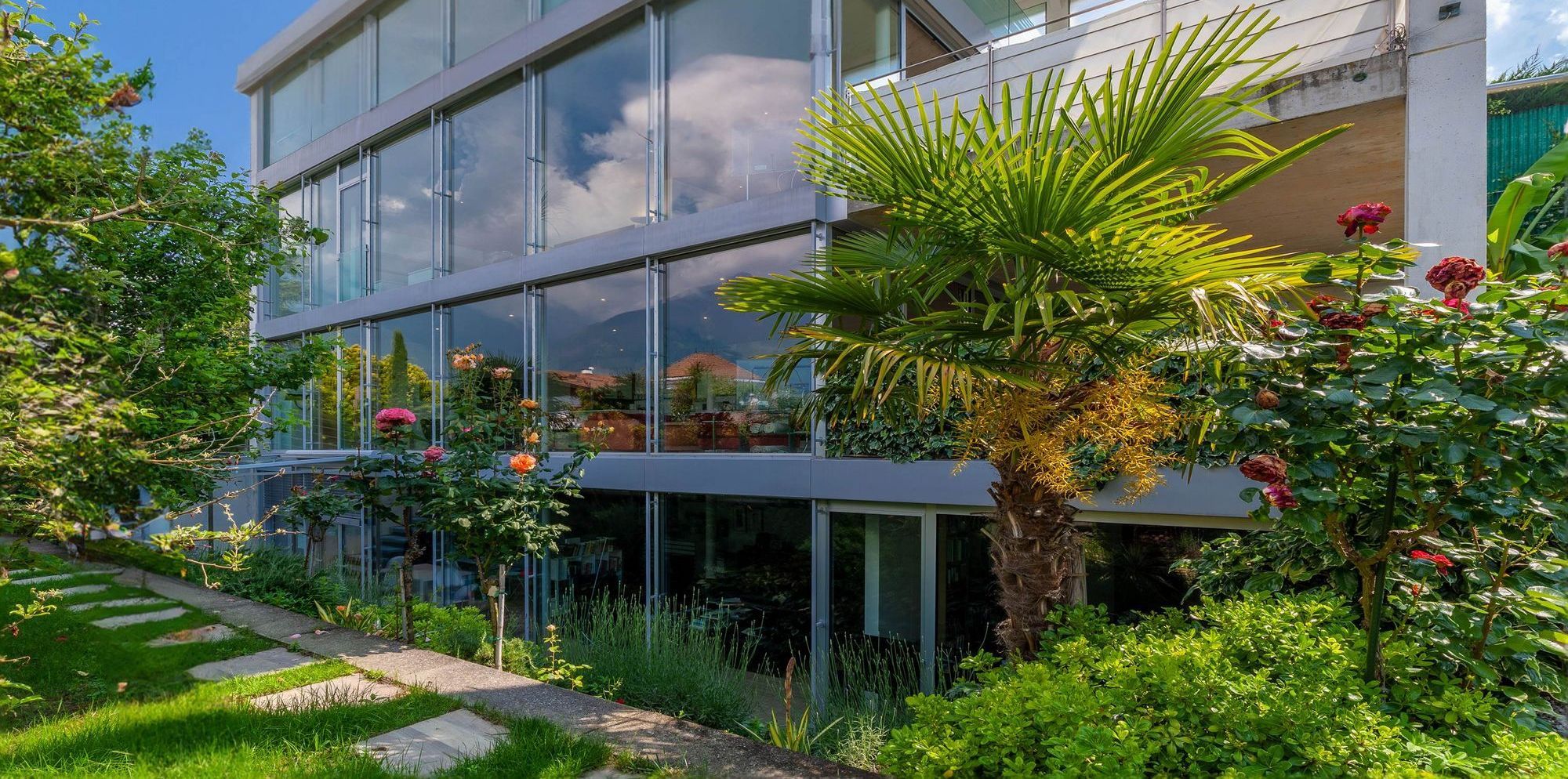 Modernes Glasgebäude mit Terrasse, Palmen und Rosen im Vorgarten