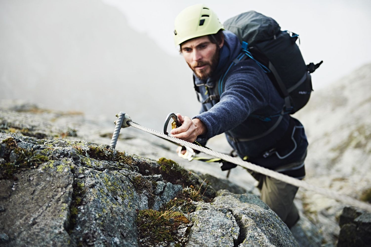 Climber with helmet and backpack clipping a carabiner to a fixed cable on a rocky slope.