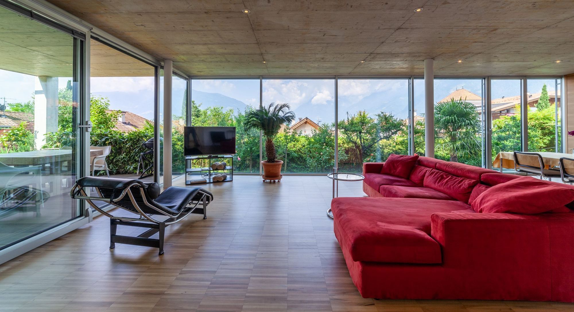 Modernes Wohnzimmer mit rotem Ecksofa, Liegestuhl und großen Glaswänden zum Garten