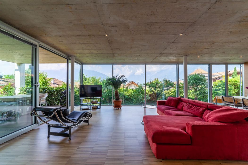 Modernes Wohnzimmer mit rotem Ecksofa, Liegestuhl und großen Glaswänden zum Garten