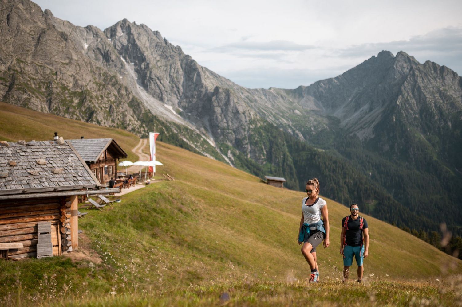 Two hikers walking on a grassy alpine slope past wooden huts with jagged mountains in the background.