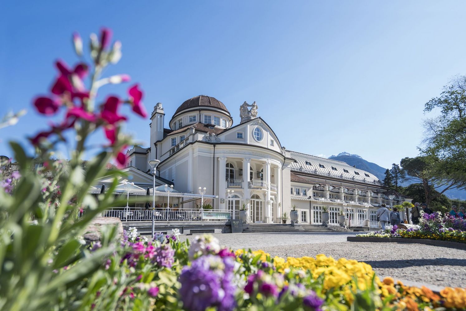 Neoclassical white spa building with a domed roof, colorful flowerbeds in the foreground and mountains under a clear blue sky.