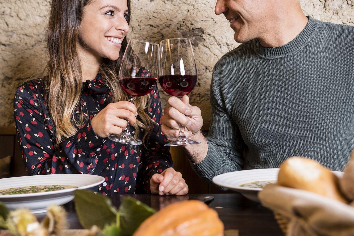 Smiling couple clinking red wine glasses over a table with soup and bread.