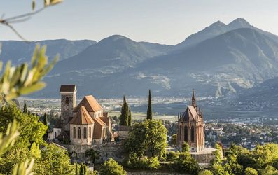 Hilltop medieval church and chapel among cypress trees overlooking a sunlit valley and distant mountains, blurred olive branches in the foreground.