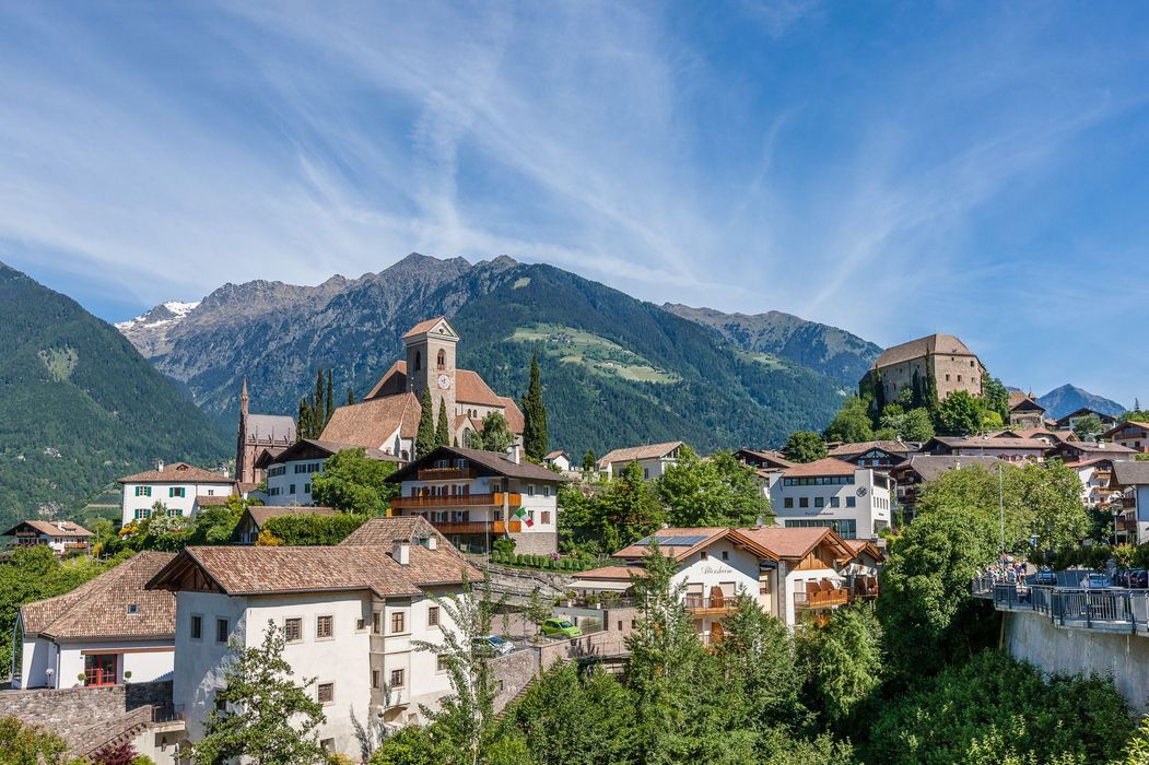 Alpenort mit Kirche und Burg vor grünen Bergen und blauem Himmel.
