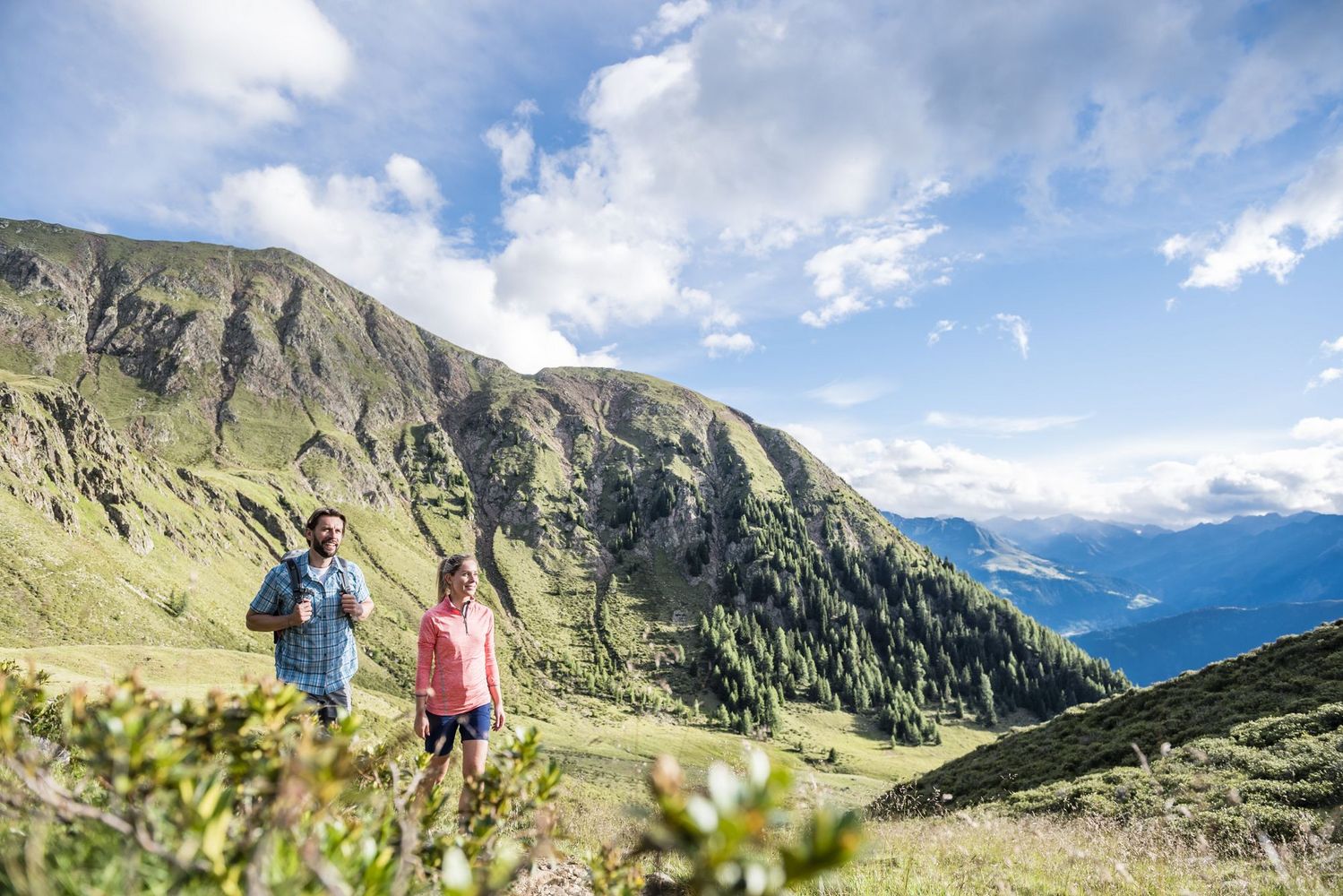 Two hikers walking on a grassy mountain trail below steep green slopes and a bright blue sky.