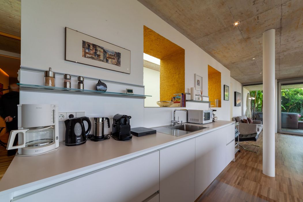 Offene moderne Küche mit weißer Arbeitsfläche, Kaffeemaschinen, Spüle und Wohnbereich im Hintergrund