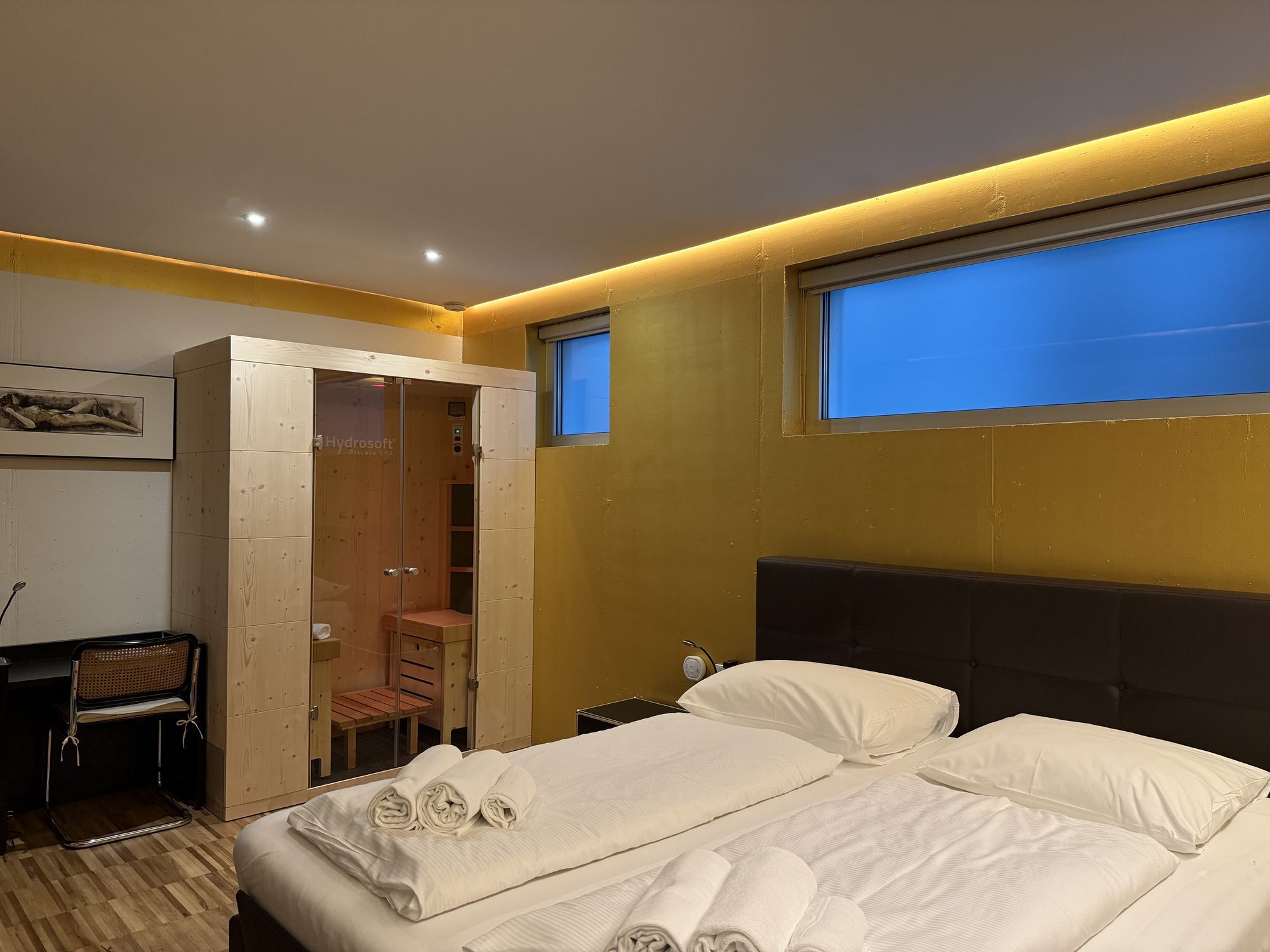 Modernes Hotelzimmer mit Doppelbett, auf dem gefaltete Handtücher liegen, und einer kleinen Holzsauna neben einem schmalen Fenster.