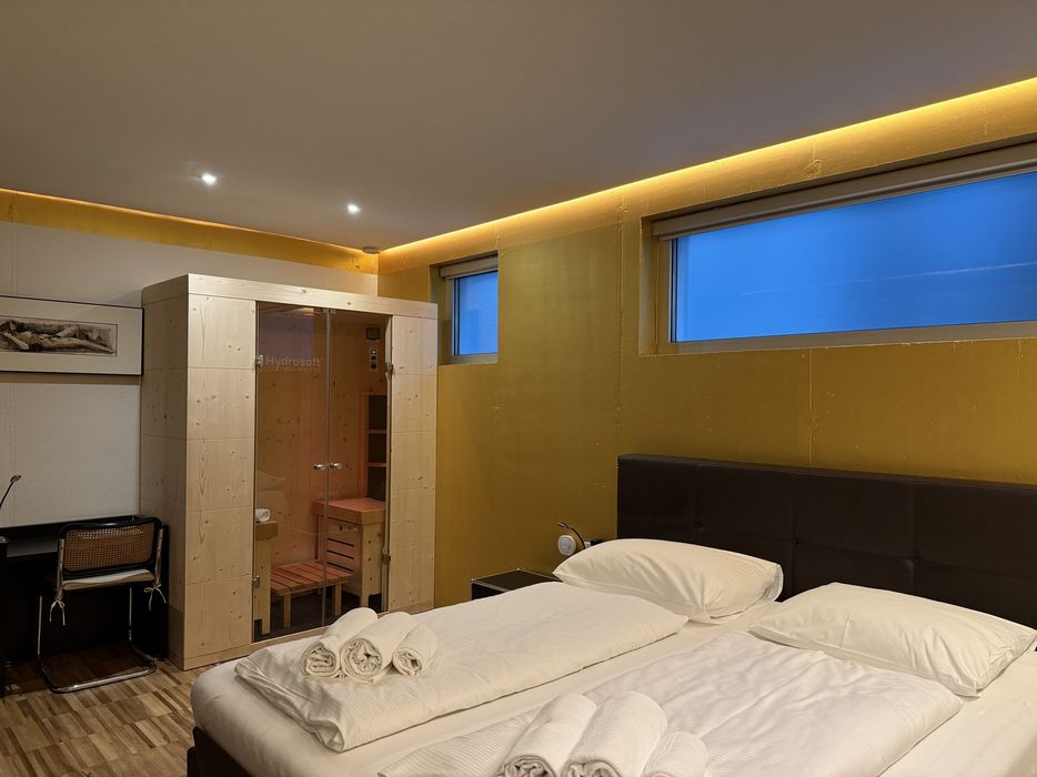 Modernes Hotelzimmer mit Doppelbett, auf dem gefaltete Handtücher liegen, und einer kleinen Holzsauna neben einem schmalen Fenster.