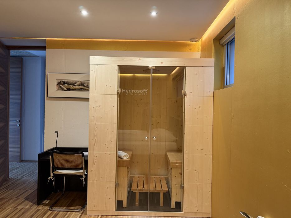 Kleine Holzsauna mit Glastür in einem Zimmer, daneben Schreibtisch und Stuhl.