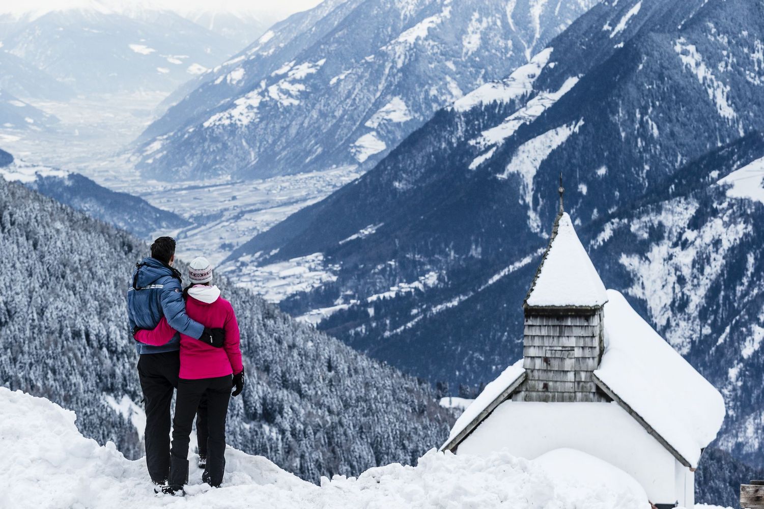 Couple in winter jackets embracing on a snowy overlook, gazing at a snow-covered valley and mountain peaks with a small chapel steeple nearby.