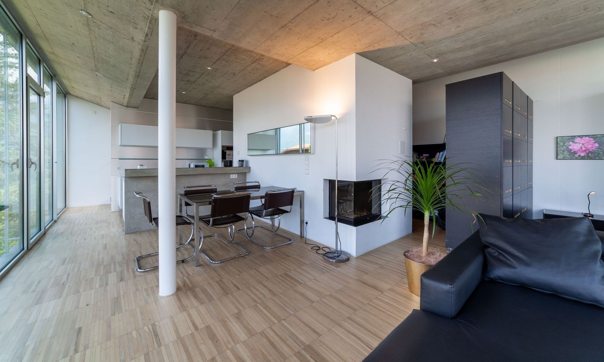 Helles, modernes Wohn- und Esszimmer mit großer Glasfront, Sichtbetondecke, Esstisch, offenem Kamin und schwarzem Sofa.