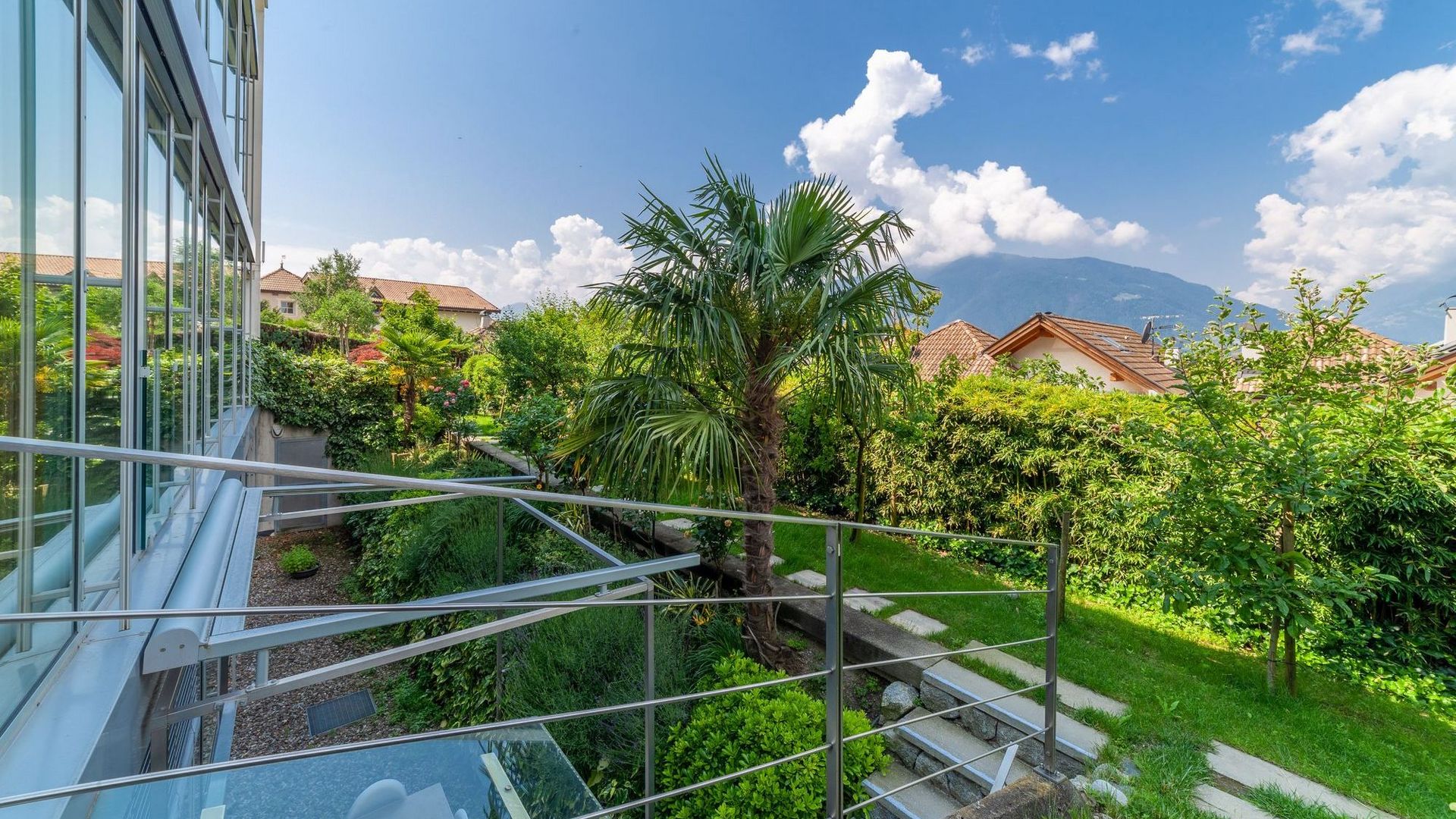 Balcone con ringhiera e facciata vetrata che guarda un giardino curato con palma, case con tetti e montagne sullo sfondo sotto un cielo azzurro.