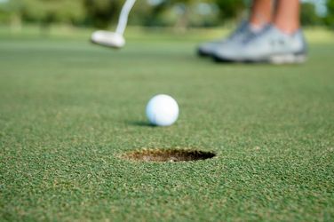 Golf ball rolling toward a hole on the putting green with a putter and shoes blurred in the background.