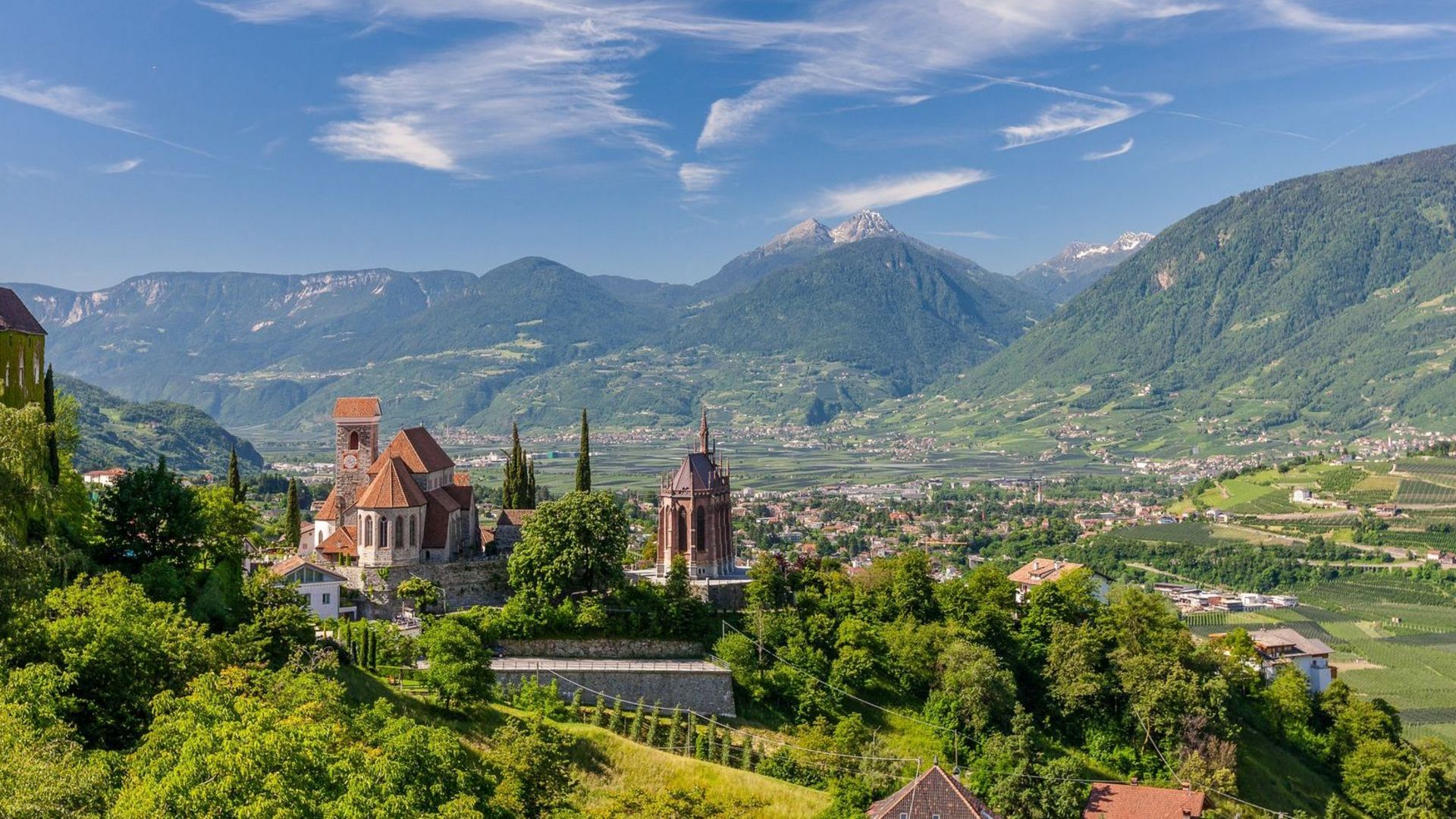 Green Alpine valley with a hillside village, church and castle beneath mountains and wispy clouds.