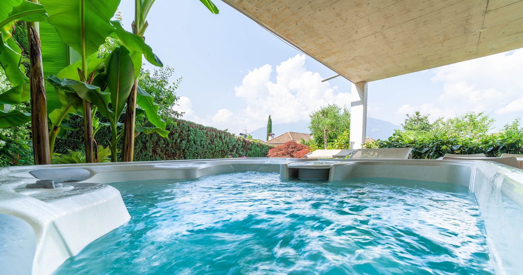 Außen-Whirlpool mit sprudelndem Wasser unter Überdachung, umgeben von Palmen und grünem Garten