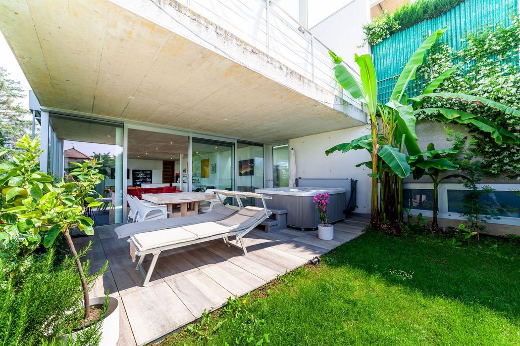 Überdachte Terrasse mit Liegestühlen, Esstisch und Whirlpool, umgeben von grünen Pflanzen und Rasen.