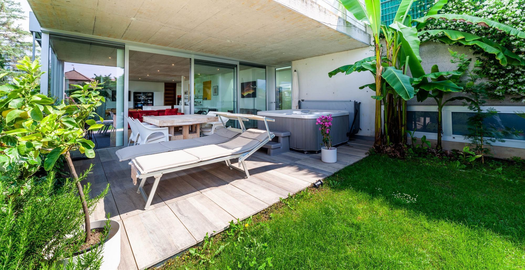 Überdachte Terrasse mit Liegestühlen, Esstisch und Whirlpool, umgeben von grünen Pflanzen und Rasen.