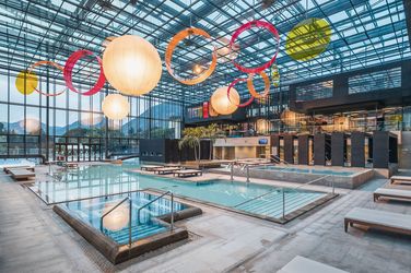 Spacious glass-roofed indoor pool complex with large hanging round lanterns and colorful circular decorations, lounge chairs beside the pools