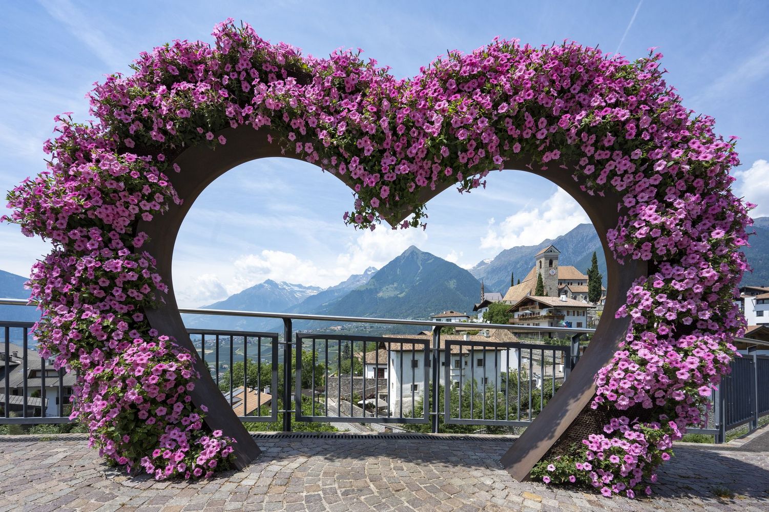 Heart-shaped frame covered in pink flowers framing a mountain village with a church and snow-capped peaks.