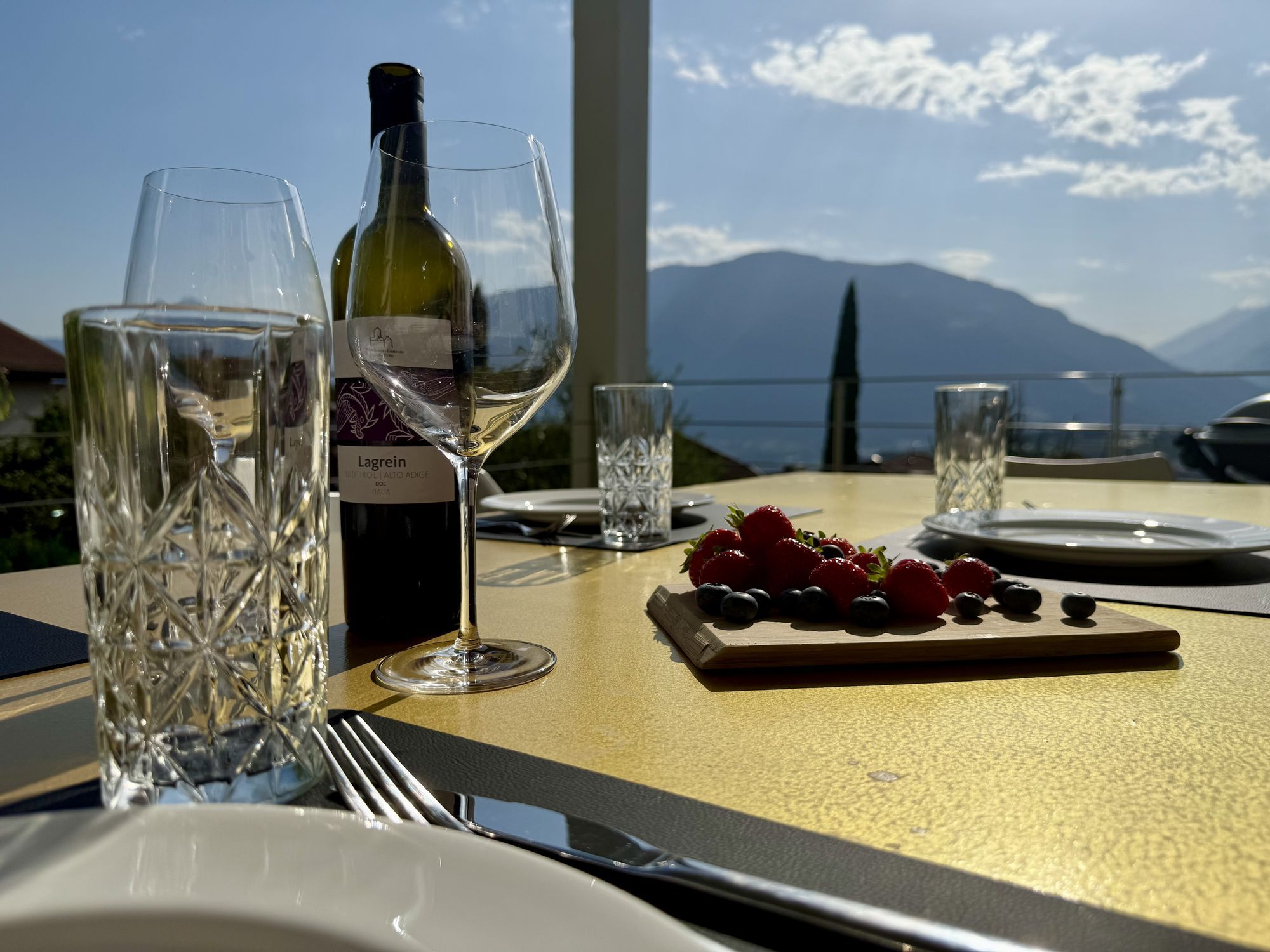 Tisch auf Terrasse mit Weinglas, Weinflasche und Brett mit Erdbeeren und Blaubeeren vor Bergpanorama
