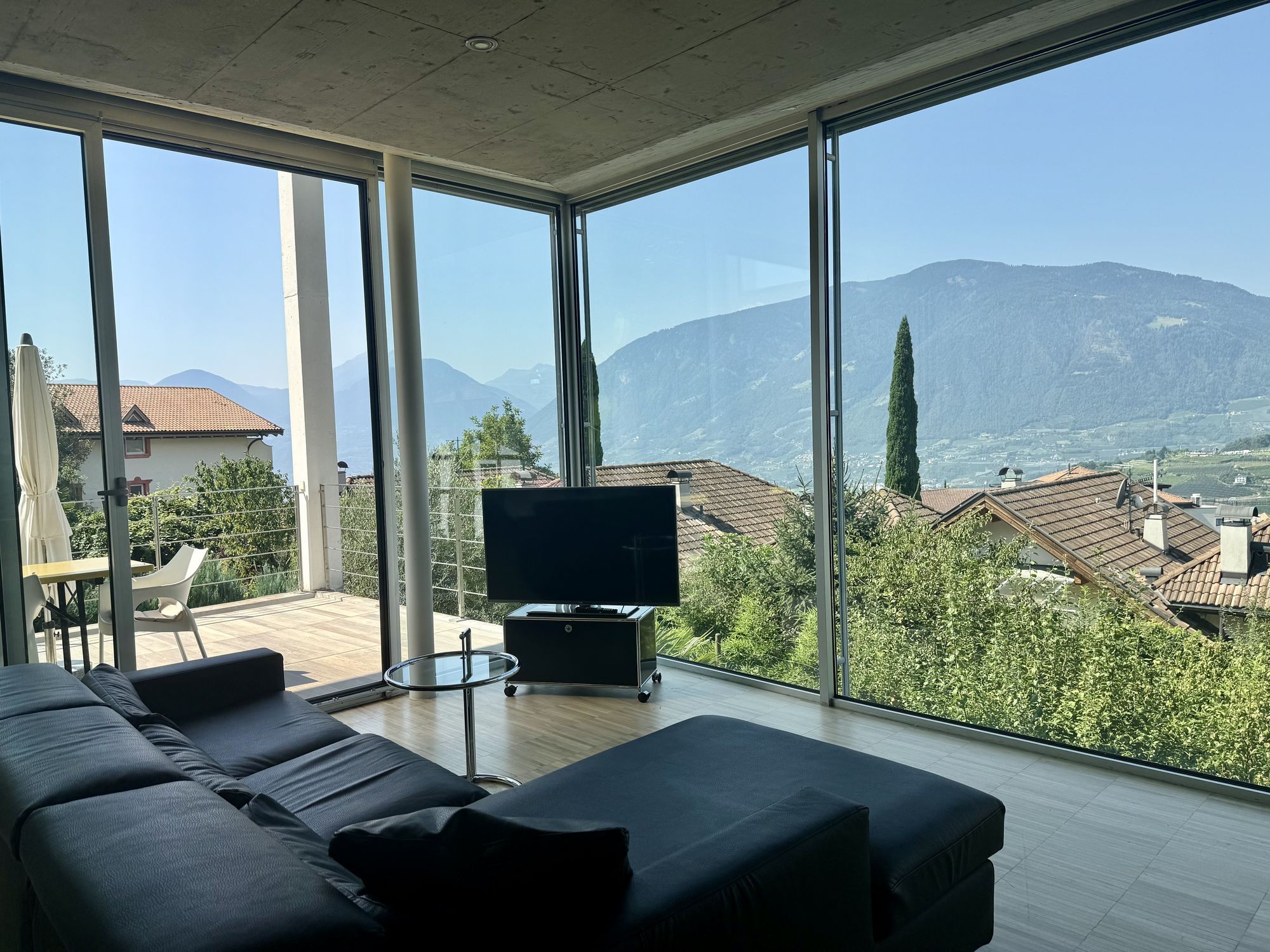 Heller Wohnraum mit schwarzem Sofa, Fernseher und bodentiefen Fenstern mit Blick auf Dächer und Berge.