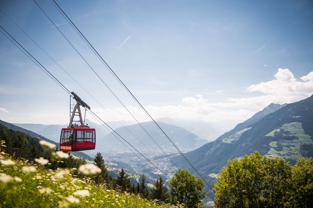 Rote Seilbahn über blühender Bergwiese mit Tal- und Alpenpanorama