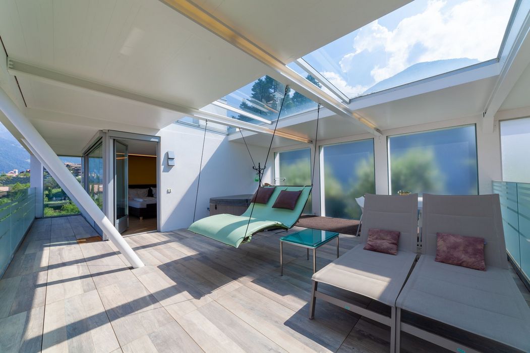 Überdachte Dachterrasse mit Hängeliege, zwei Sonnenliegen und Glasdach mit Bergblick.