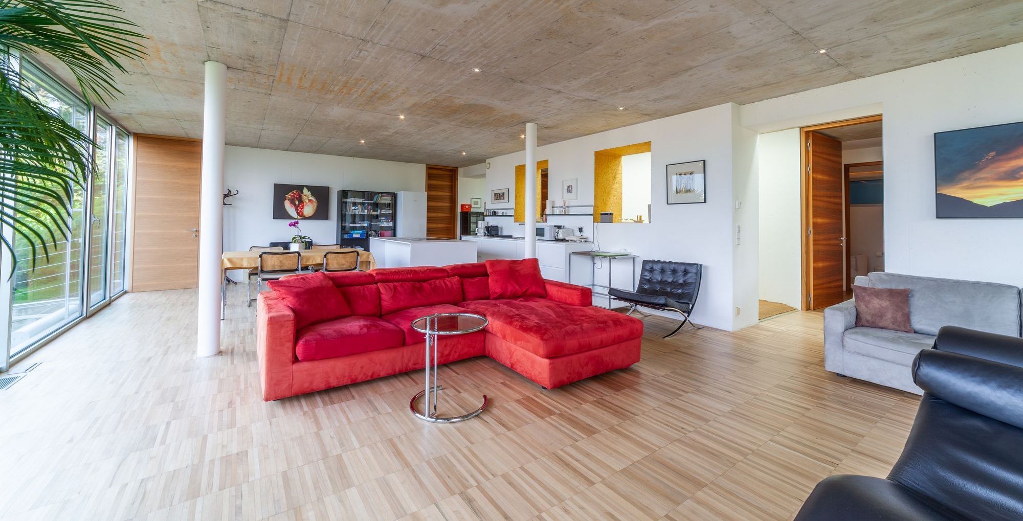 Moderner offener Wohnraum mit rotem Ecksofa, großen Panoramafenstern, Beton­decke und Holzfußboden.