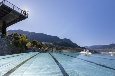 Two people on a viewing platform above a sunlit outdoor pool with mountains in the background.