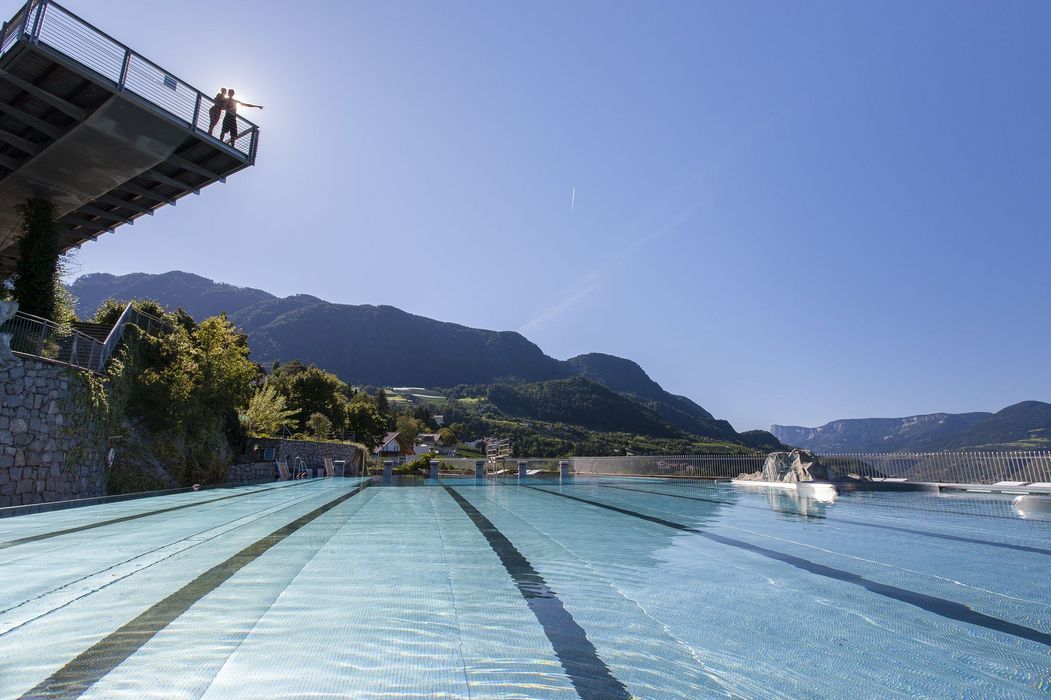 Freiluft-Schwimmbecken mit Bahnen vor Bergen, zwei Personen auf Aussichtsplattform