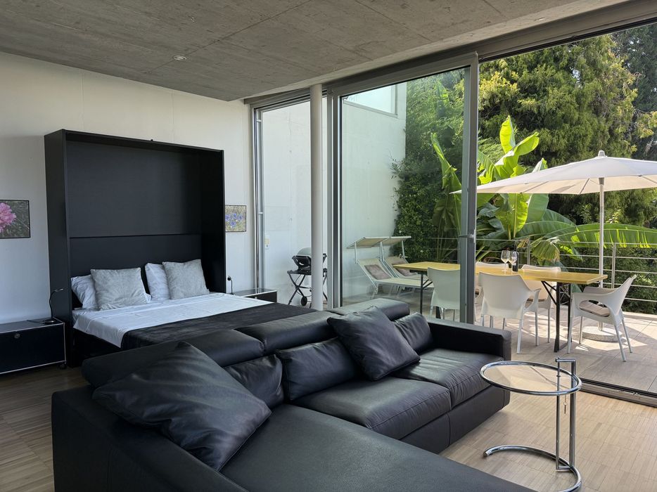 Studio mit schwarzem Sofa und Bett, große Glas-Schiebetür zum Balkon mit Tisch, Sonnenschirm und Pflanzen.