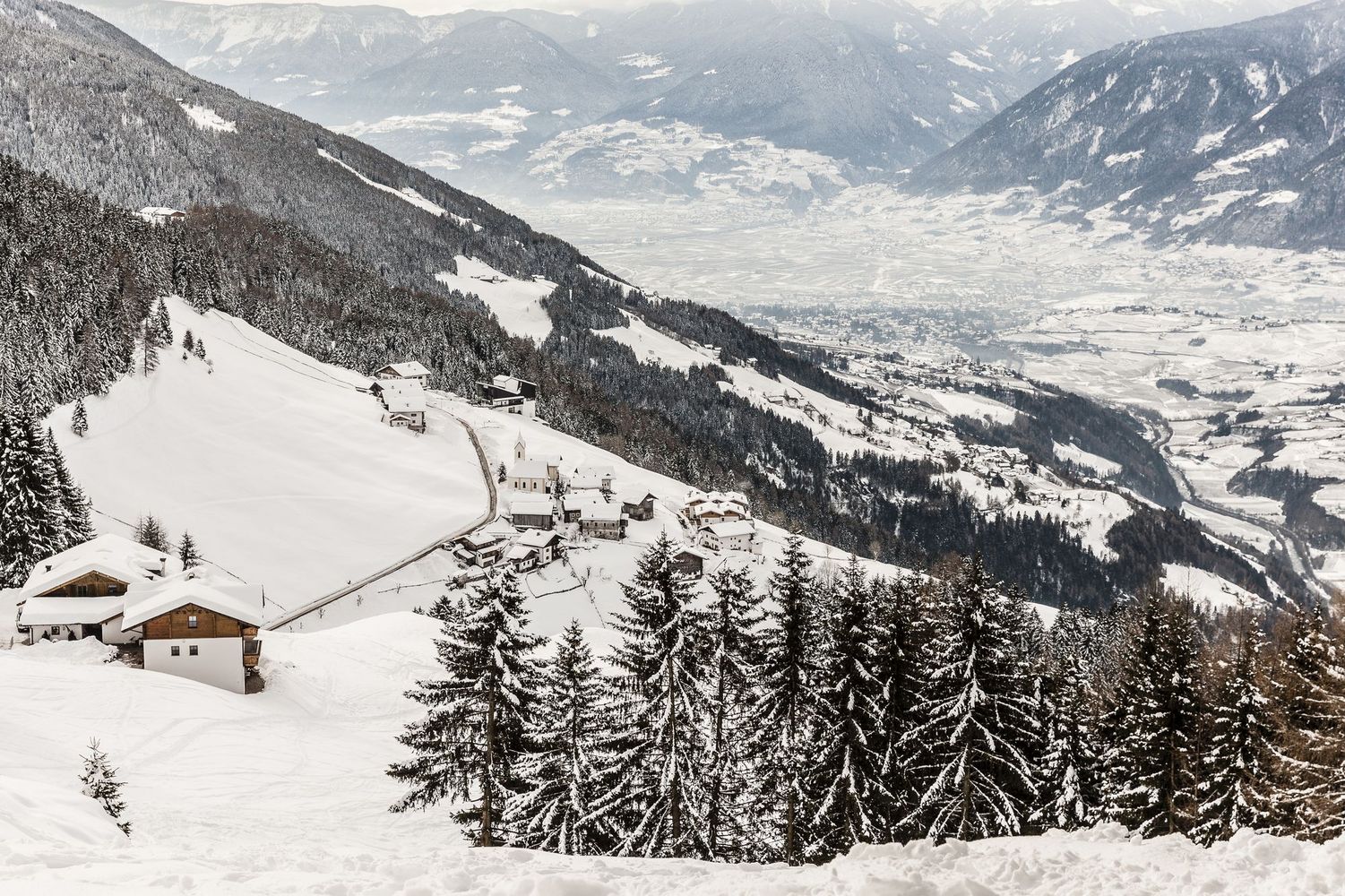 Snow-covered alpine village on a mountainside with fir trees and a distant valley.