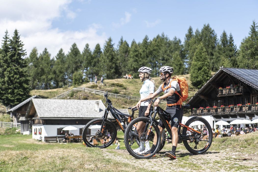 Zwei Radfahrer mit Helmen neben Mountainbikes vor Almhaus und Nadelwald
