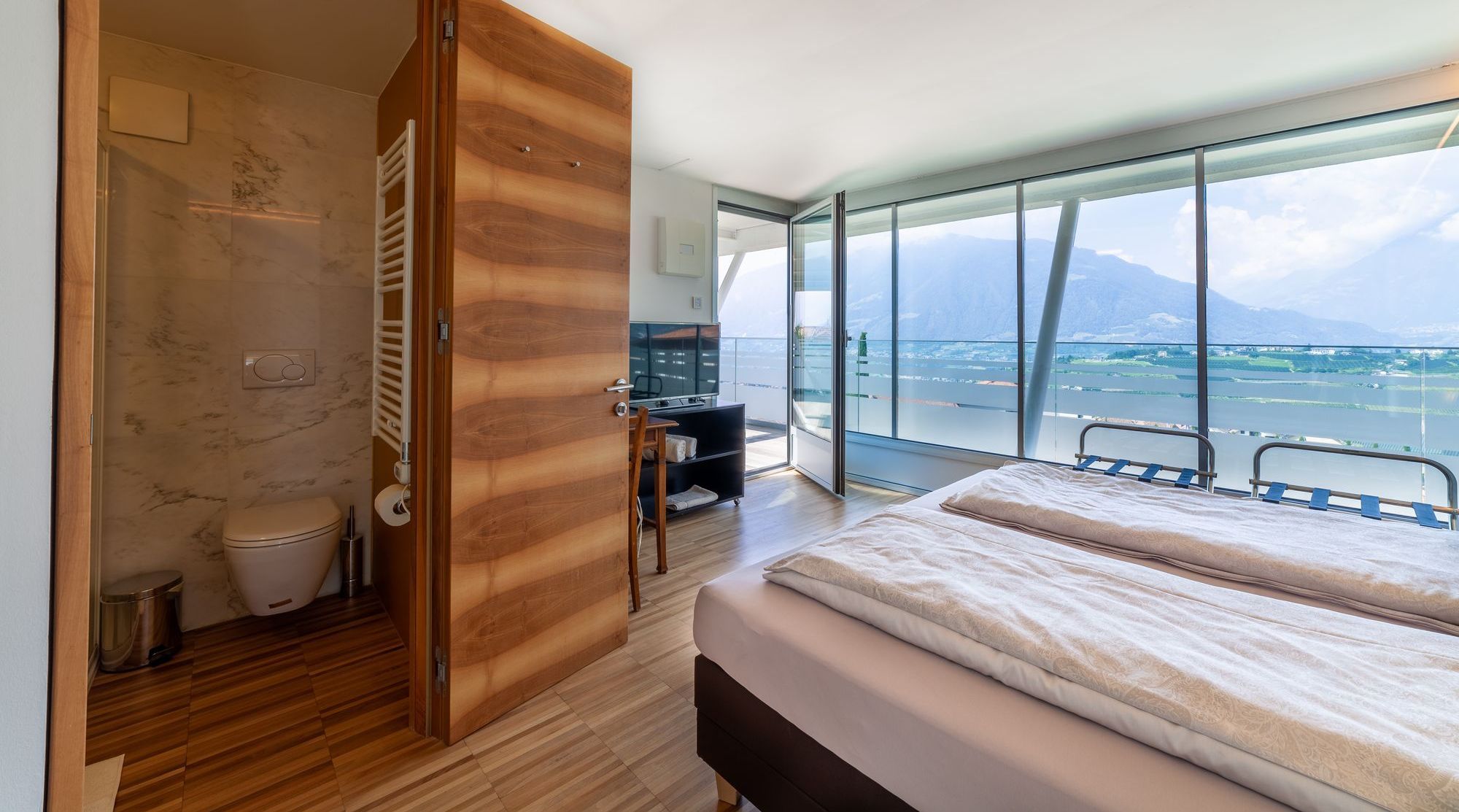 Schlafzimmer mit Doppelbett, offenem Bad und Panoramafenstern mit See- und Bergblick