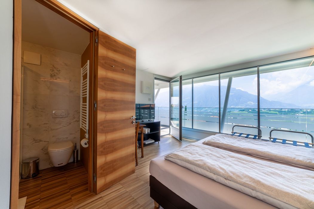 Schlafzimmer mit Doppelbett, offenem Bad und Panoramafenstern mit See- und Bergblick