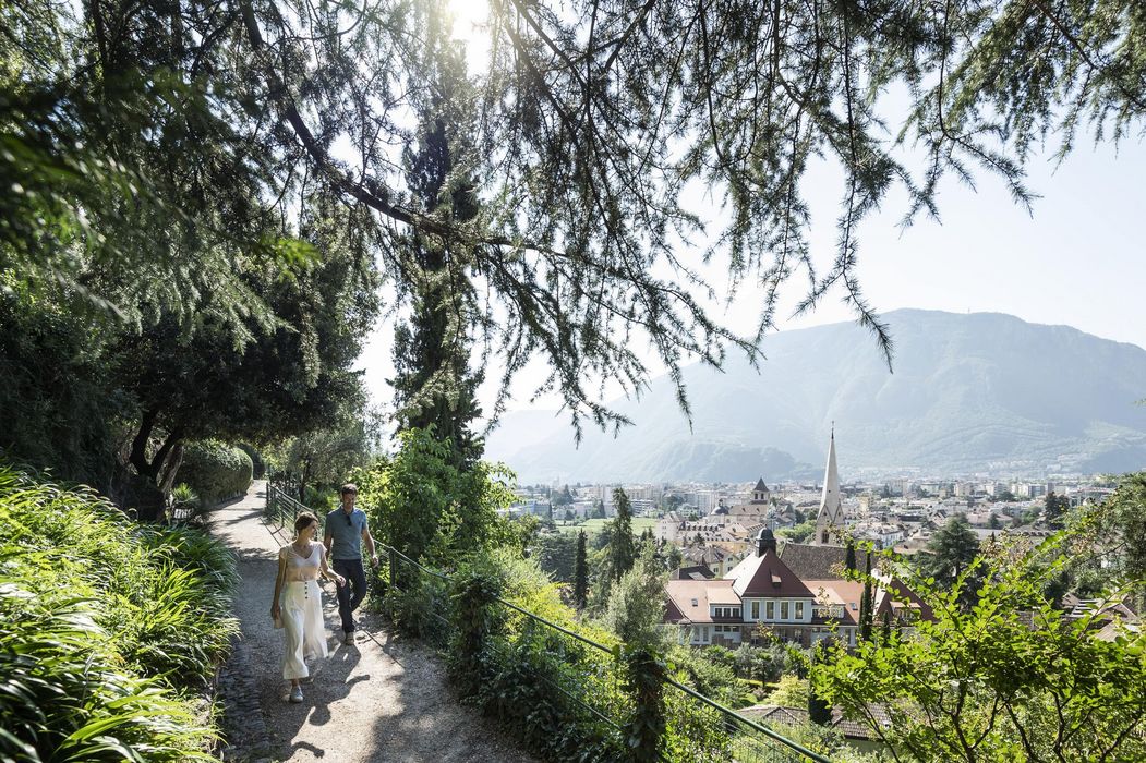 Paar spaziert auf schattigem Hangweg mit Blick auf Stadt, Kirchturm und Berge