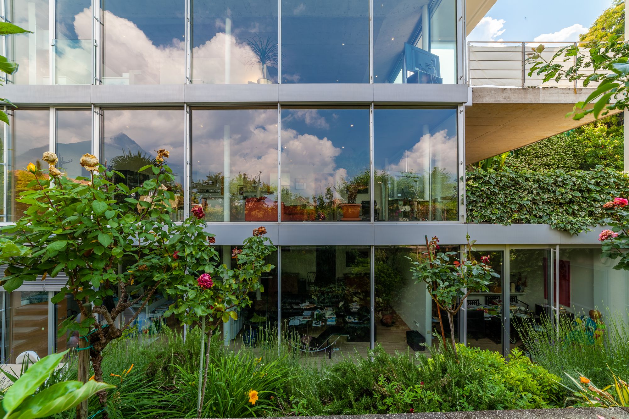 Facciata vetrata di una casa moderna che riflette nuvole e montagne, con rose e giardino in primo piano.