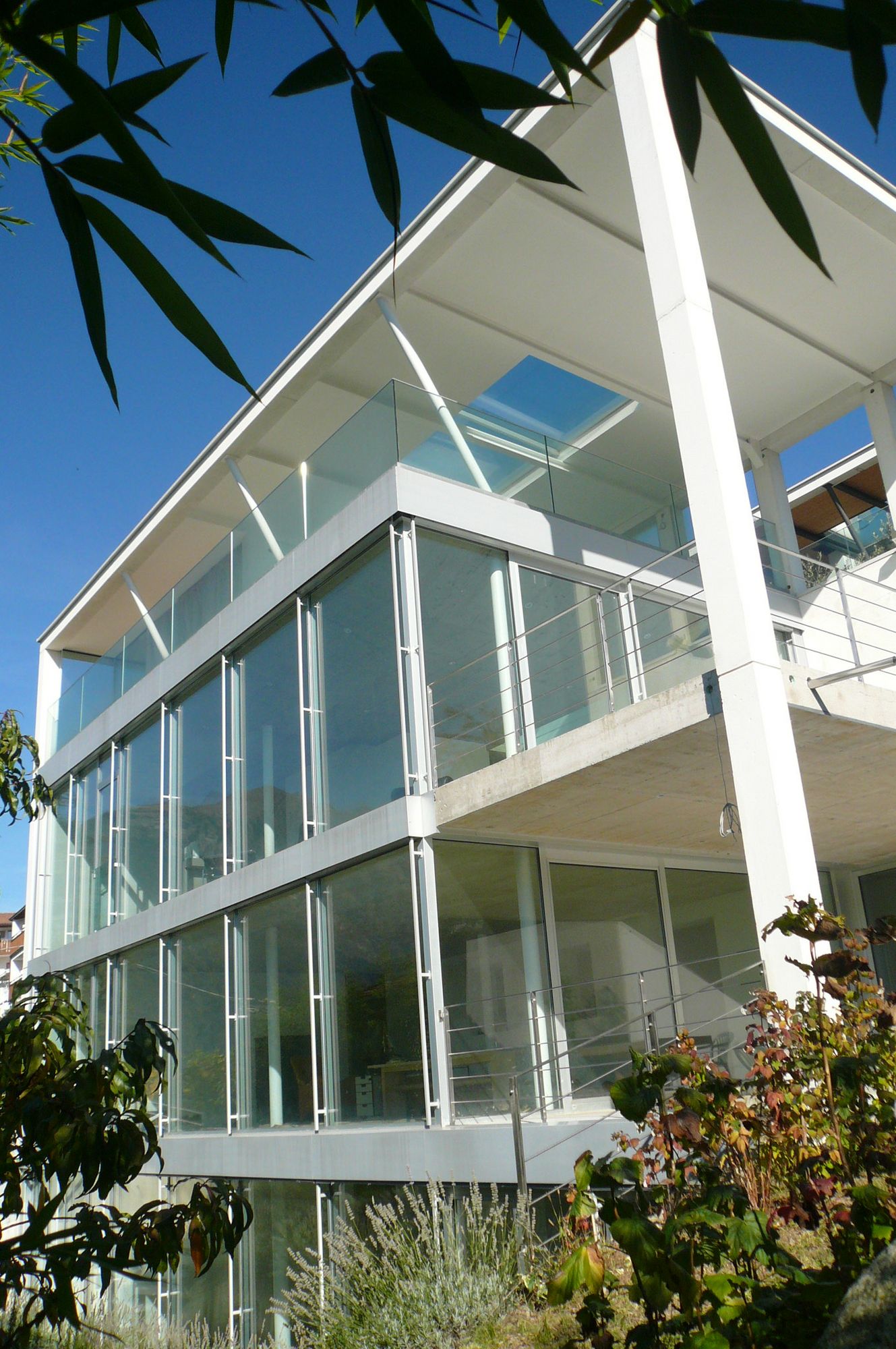 Casa moderna bianca a più piani con ampie vetrate, balconi, cielo azzurro e piante in primo piano.