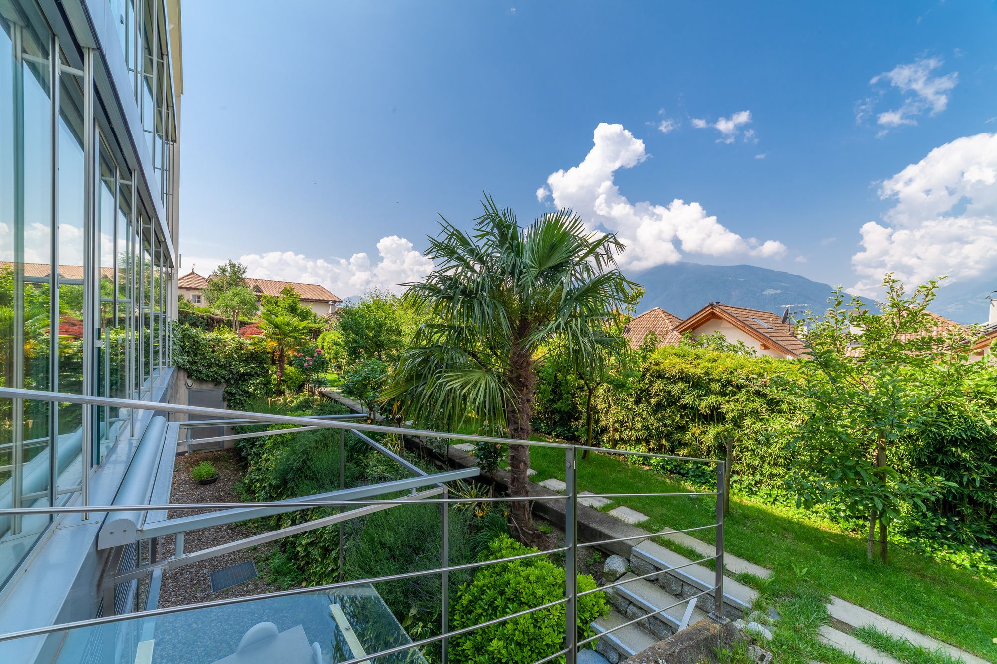 Balcone con ringhiera e facciata vetrata che guarda un giardino curato con palma, case con tetti e montagne sullo sfondo sotto un cielo azzurro.
