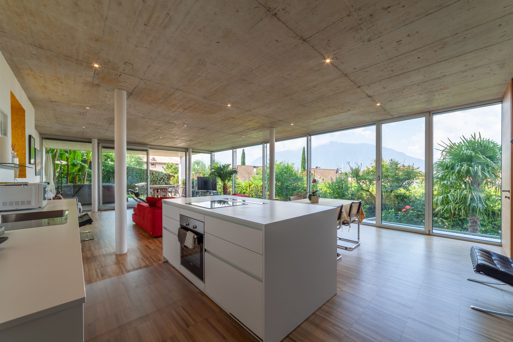 Open-space con cucina a isola, divano rosso e grandi vetrate sul giardino e le montagne