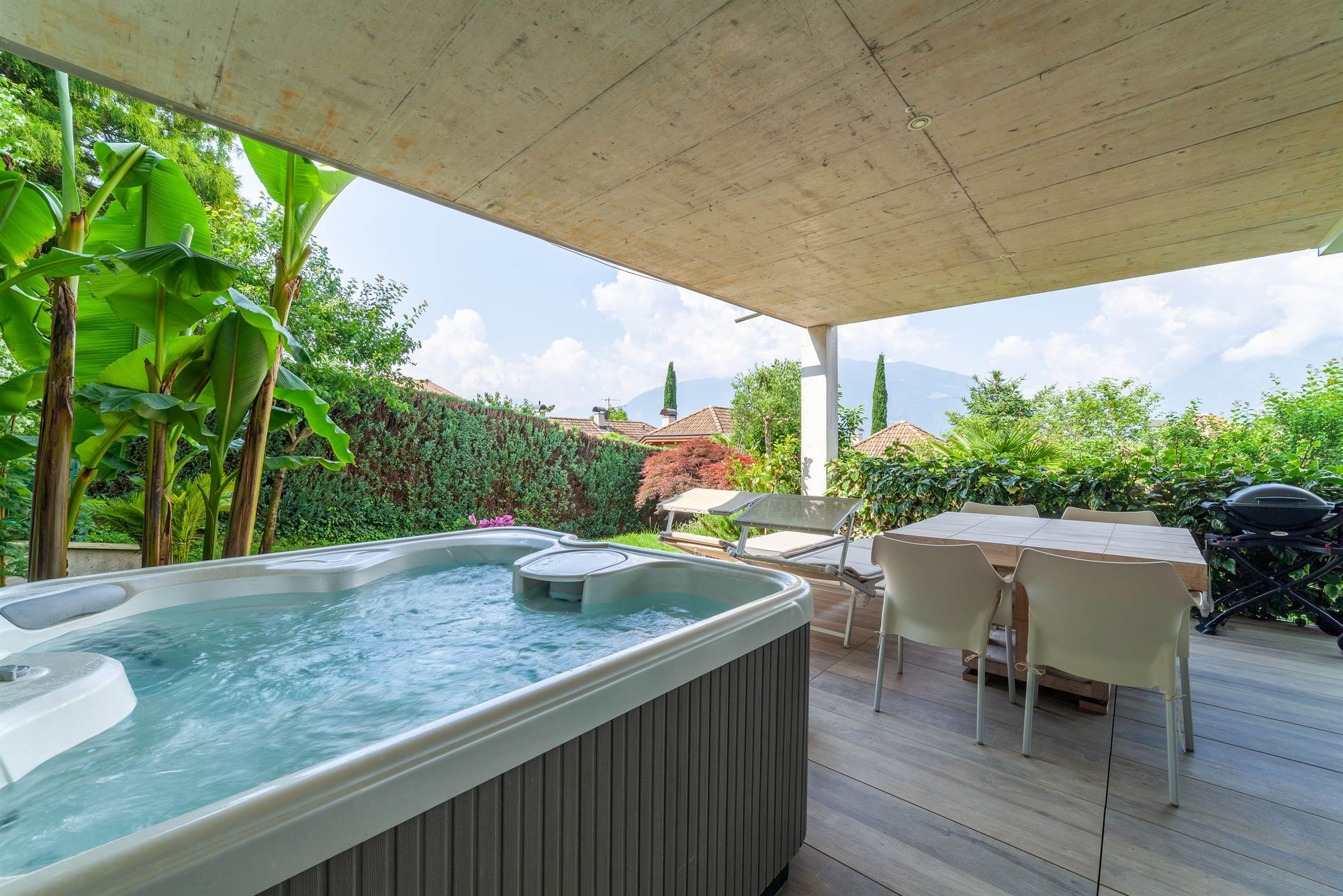 Veranda coperta con vasca idromassaggio, tavolo con sedie e lettini, giardino rigoglioso e vista sulle colline.